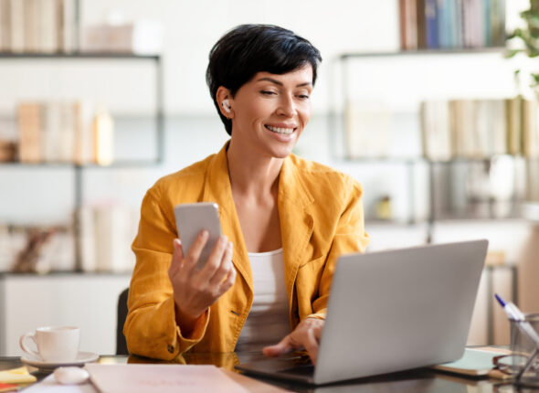 Businesswoman with earbuds and phone working on laptop in office